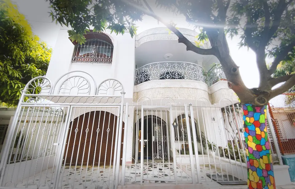 La casa vista desde la calle: dos pisos blancos, balcón curvo de hierro forjado, rejas con abanicos y el árbol de tronco pintado