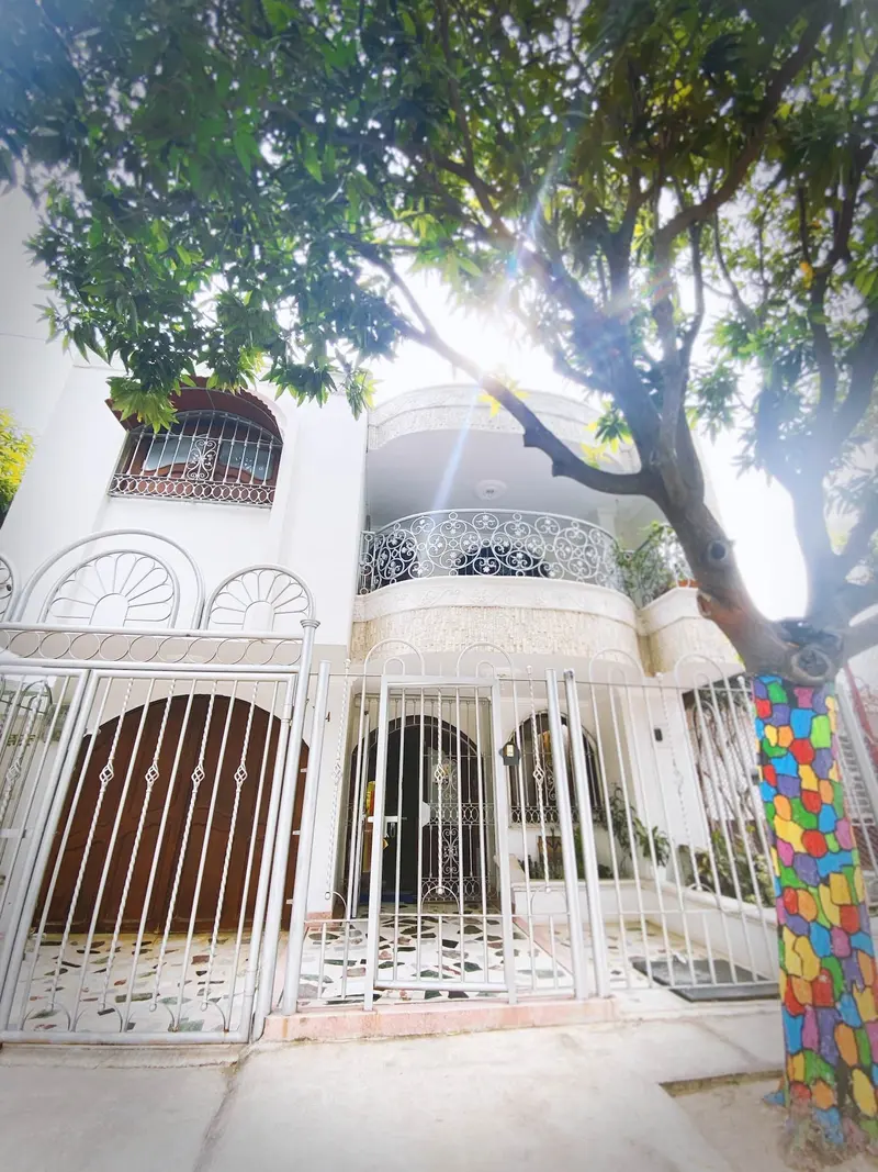Fachada blanca de Tía Chela: balcón curvo con baranda de hierro forjado, rejas en arco y el sol colándose entre las ramas del árbol de la entrada